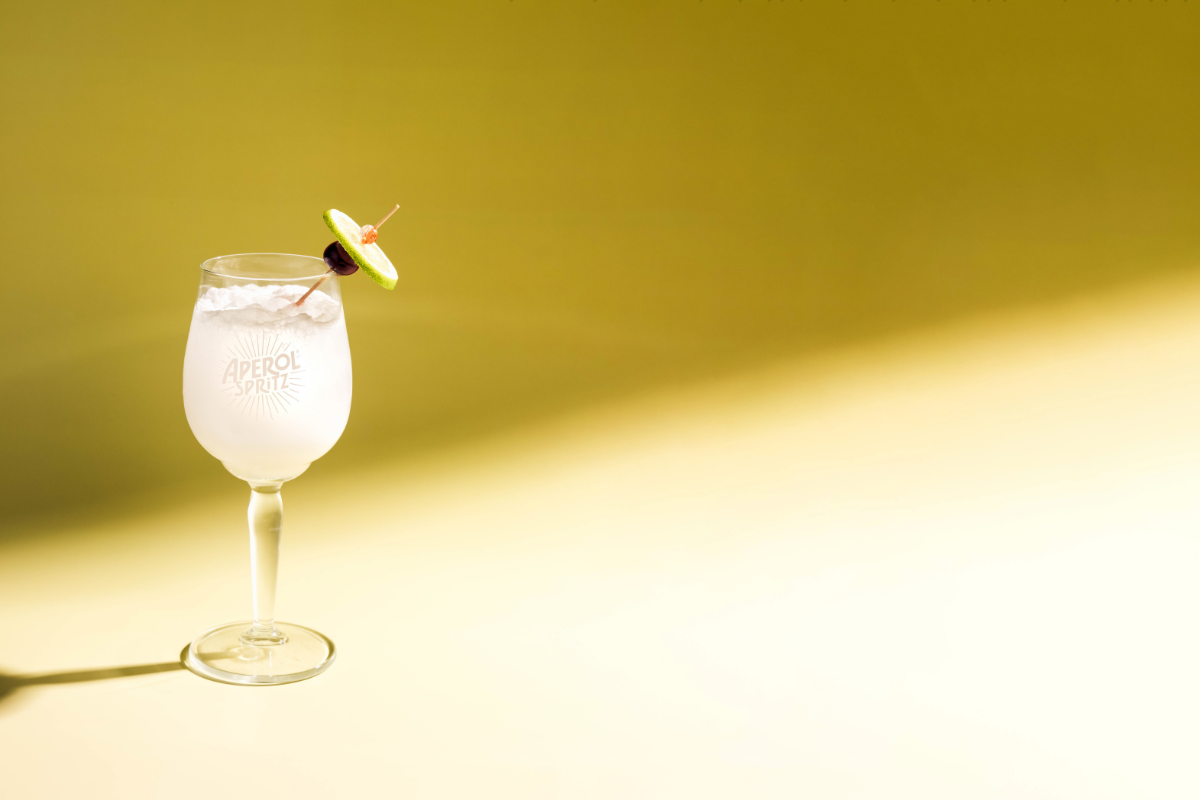 Coco Blanco Spritz from Punch Bowl Social, a light coconut cocktail served over ice in an Aperol glass with a lime and cherry garnish on a yellow backdrop.
