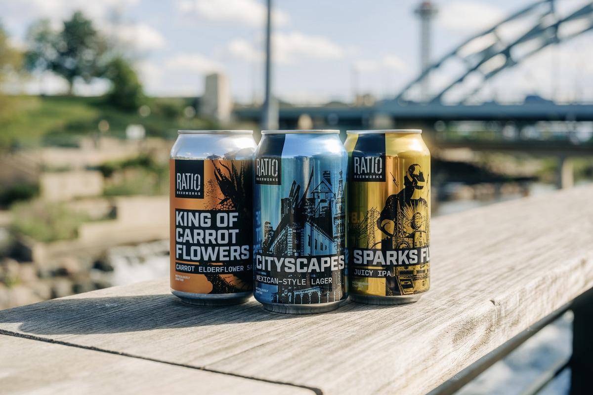 Ratio_Beerworks Three Ratio Beerworks cans—King of Carrot Flowers, Cityscapes, and Sparks Fly—displayed outdoors on wood railing with bridge and skyline backdrop.