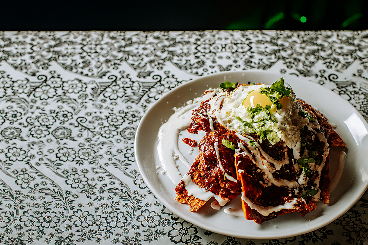 Red Mole Chilaquiles_Photo courtesy of La Diabla Pozole y Mezcal A plate of red mole chilaquiles topped with a runny fried egg, crema, crumbled cheese, and fresh cilantro sits on a white plate over a black-and-white floral tablecloth, lit from the side with soft, warm light.