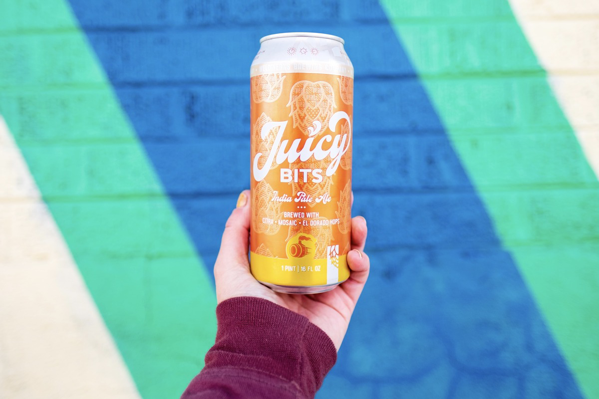 Weldwerks_Brewing_Co Hand holding a Juicy Bits IPA from WeldWerks Brewing Co. in front of a colorful mural, showcasing the hazy beer brewed with Citra, Mosaic, and El Dorado hops.