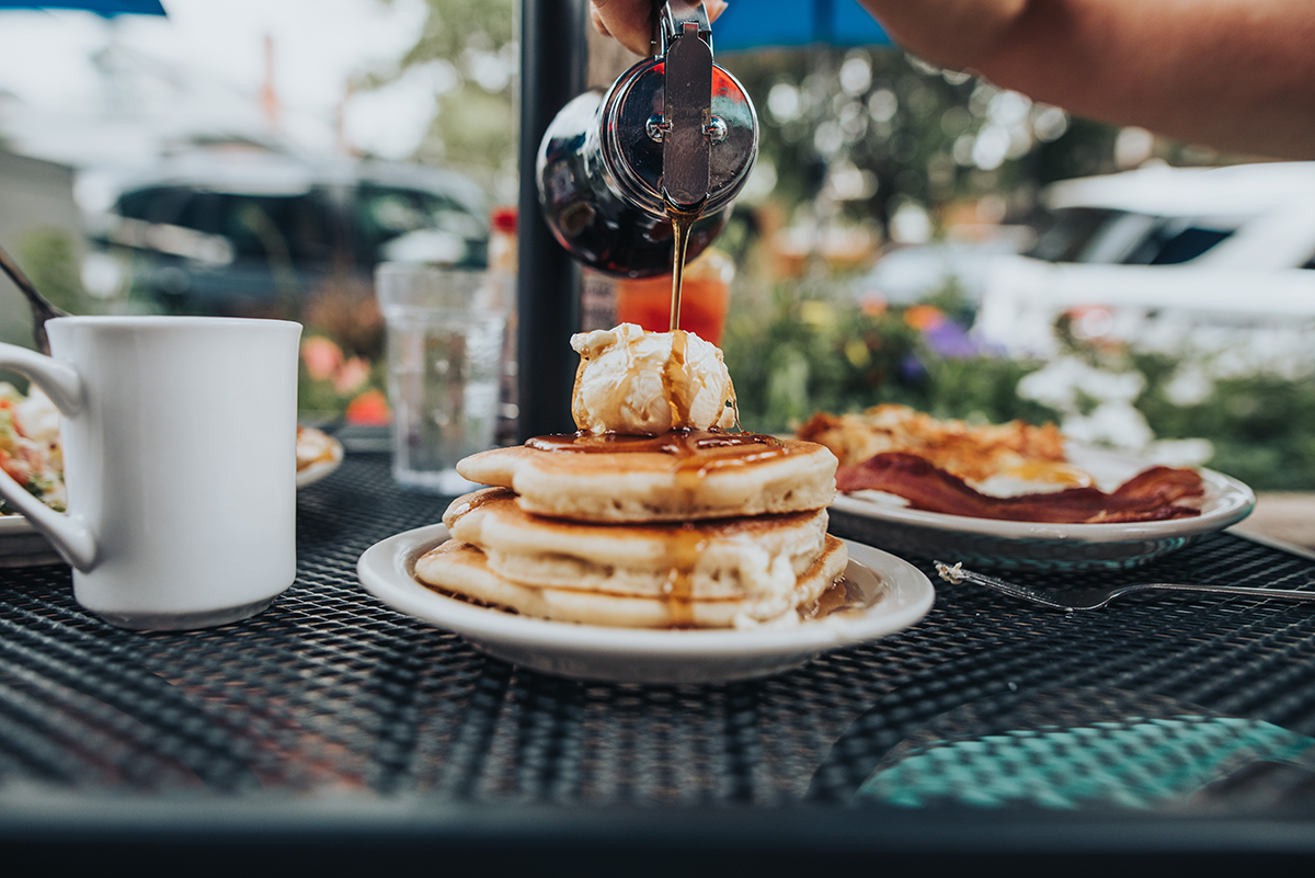 zorbas all-day breakfast A hand pours syrup over a stack of fluffy pancakes topped with a scoop of butter or whipped cream, with bacon and eggs on a plate in the background on an outdoor patio table.