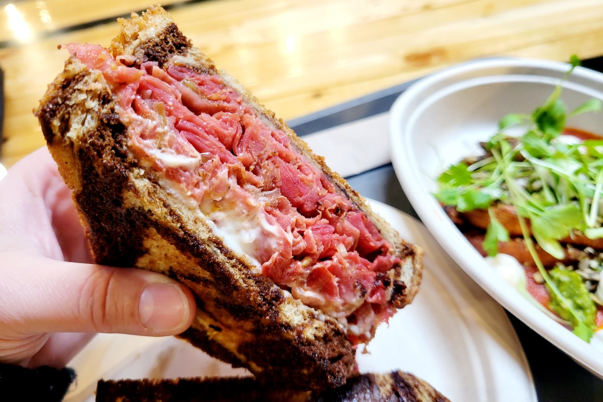 A hand holds up a thick Reuben sandwich packed with corned beef and melty cheese on toasted rye at Gladys in Denver, with fries and greens in the background.