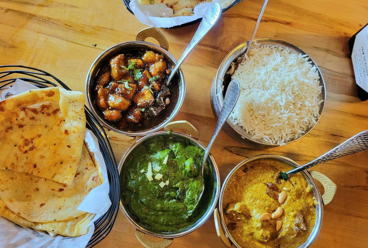 A colorful Indian meal at Spice Room in Denver, featuring naan, basmati rice, palak paneer, korma, and Manchurian in metal bowls on a wooden table.
