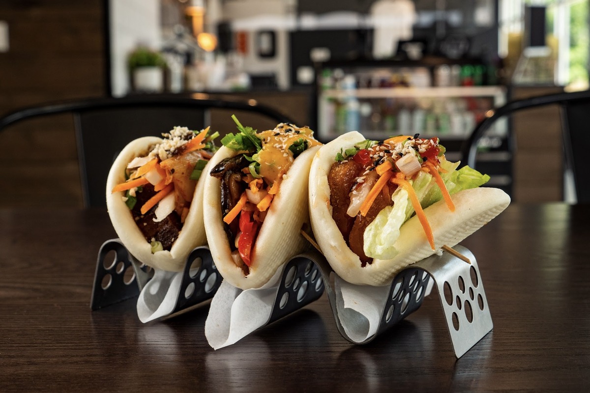 Trio of vegan bao buns from V Revolution in Denver, filled with crispy tofu, veggies, fresh herbs, and topped with sauces and sesame seeds on a metal stand.
